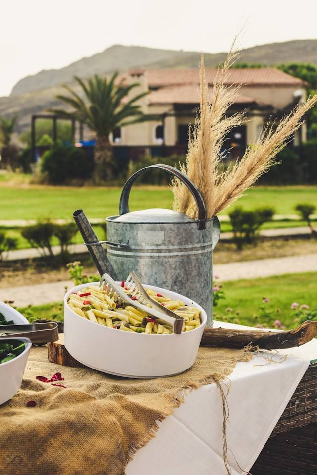Eine Schüssel Nudelsalat auf einem Tisch im Freien, dahinter ein mediterranes Haus mit Ziegeldach und Palmen